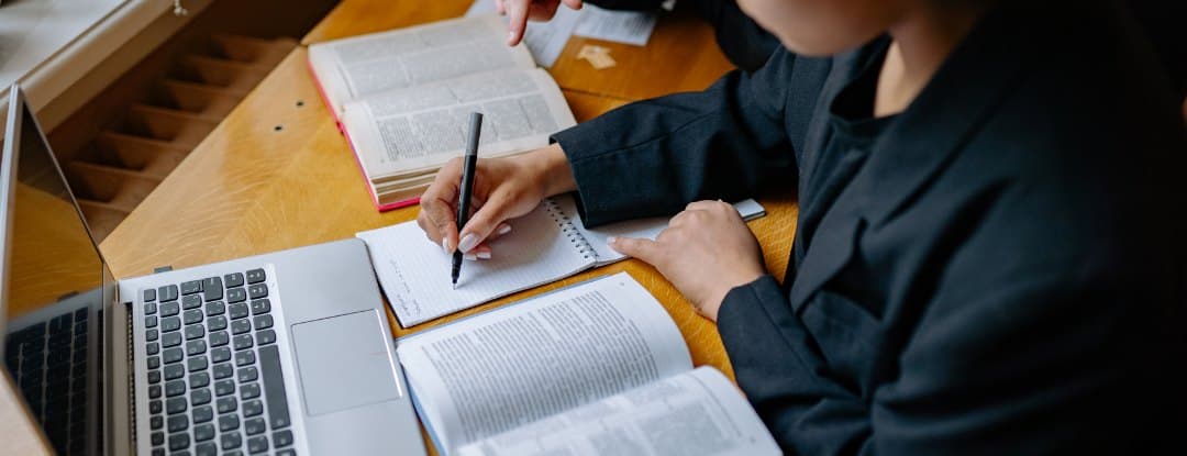 Studentka u notebooku píše poznámky do sešitu, kolem otevřené učebnice